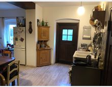 Gemütliche Küche mit Holzelementen: ein Holzfußboden, ein dunkler Holztürrahmen sowie eine einfache Holzeinrichtung. Ein großer Kühlschrank steht neben einer Eckkommode, über der sich Regale mit Küchenutensilien befinden. Links ein Fenster mit weißen Vorhängen, rechts eine kleine Spüle und Kochfläche.