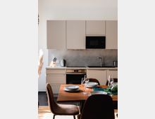 Moderne Küche mit hellgrauen Schränken, einem eingebauten Backofen, einer Mikrowelle und einem Wasserhahn vor einer grauen Wandverkleidung. Im Vordergrund steht ein gedeckter Holztisch mit Tellern, Gläsern und braunen Stühlen.