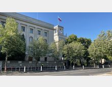 Großes historisches Gebäude mit heller Fassade und Säulen, flankiert von mehreren Bäumen. Auf dem Dach weht eine russische Flagge, und vor dem Gebäude befinden sich Absperrungen entlang der Straße.