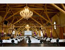 Großer Veranstaltungssaal in einer rustikalen Scheune mit sichtbarer Holzbalkendecke. Der Raum ist mit Kronleuchtern, Lichterketten, weiß gedeckten Tischen und schwarzen Stühlen ausgestattet, im Hintergrund ist eine Leinwand zu sehen.