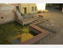 Historischer Innenhof mit gepflastertem Boden, einem kleinen Wassergraben mit Algenbewuchs und Backsteinmauern. Eine Treppe führt zu einem Eingang, daneben Sitzbänke und Geländer entlang des Geländes.