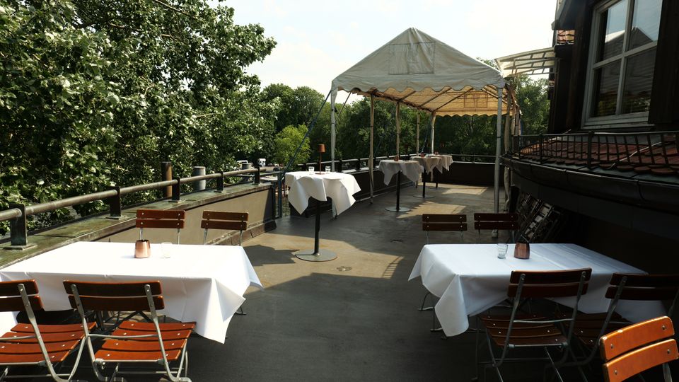 Dachterrasse eines Restaurants mit mehreren Tischen mit weißen Tischdecken und Holzklappstühlen, teilweise unter einem weißen Pavillonzelt. Links grenzt ein Geländer an dichtes Grün, rechts verläuft die Fassade des Gebäudes mit Fenstern.