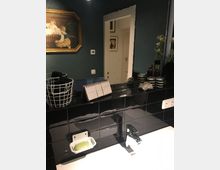 Waschraum mit dunkelgrünen Wänden und schwarzen glänzenden Fliesen an der Wand. Ein weißes Waschbecken mit verchromtem Wasserhahn und passender Seifenschale steht im Vordergrund, während Dekorartikel, ein Bilderrahmen und ein Wandspiegel den Raum ergänzen.