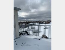 Schneebedeckte Dachterrasse mit Geländer, umliegenden Dachfenstern und teilweise wolkenverhangenem Himmel; im Hintergrund sind städtische Gebäude und Schornsteine sichtbar.