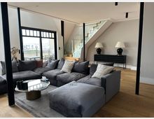 Modernes Wohnzimmer mit großer grauer Eckcouch auf Teppich und rundem Couchtisch vor einer breiten Glasfront mit Zugang zur Terrasse. Im Hintergrund führt eine Treppe mit Holzstufen und Glasgeländer nach oben; daneben stehen ein Sideboard mit zwei Tischlampen und schwarze Stützpfeiler im Raum.