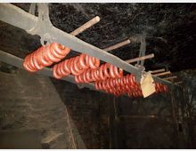 Räucherraum mit an Eisenstangen aufgehängten, ringförmigen Würsten; dunkle, rußgeschwärzte Wände und Decke.
