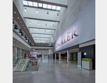 Großzügiger, moderner Innenbereich eines Büro- oder Ausstellungsgebäudes mit grauem Betonwänden, großen Fenstern und einem offenen Treppenaufgang. An der rechten Wand ist ein großes Firmenlogo mit „Daimler“ angebracht; die Decke ist mit mehreren hängenden Leuchten ausgestattet, und im Hintergrund sind Pflanzen und ein Fahrzeugausstellungsbereich sichtbar.