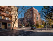 Straßenszene in einer Stadt mit gelbem Bus an der Kreuzung, umgeben von mehrstöckigen Gebäuden aus Ziegel und verputztem Mauerwerk, kahlen Bäumen und Ampeln im Vordergrund. Ein herbstlicher Tag mit blauem Himmel und vereinzeltem Straßenverkehr.