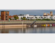 Uferansicht mit moderner Wohnbebauung und einem historischen Backsteingebäude im Hintergrund. Im Vordergrund befindet sich ein Hafenbereich mit einer Anlegestelle, Booten und bunten Tretbooten auf dem Wasser.