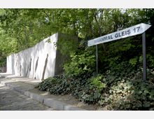 Gedenkort mit einer Betonwand, in die Silhouetten von Menschen eingearbeitet sind, flankiert von dicht bewachsenen Bäumen und einem Gehweg mit Kopfsteinpflaster; ein Schild mit der Aufschrift „Mahnmal Gleis 17“ steht rechts im Bild.