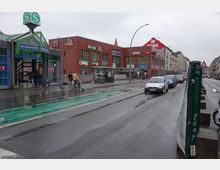 Straßenszene in einer urbanen Umgebung mit dem Eingang zur S-Bahn-Station "Hermannstraße" auf der linken Seite und einem Backsteingebäude mit Geschäften wie DM und Rewe im Hintergrund. Fahrräder stehen am Gehweg, während Autos die nasse Straße entlangfahren.