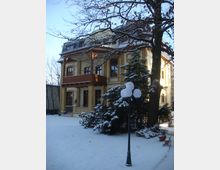 Wohnhaus im historischen Stil mit gelben Klinkerfassaden und Holzveranda, umgeben von verschneitem Garten mit Bäumen und einer Straßenlaterne im Vordergrund. Die Umgebung wirkt winterlich mit Schnee bedecktem Boden und Ästen.