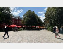 Platz mit Kopfsteinpflaster vor einem historischen roten Backsteingebäude. Im Vordergrund stehen Bäume und rot-gelbe Sonnenschirme, die zu einem Außenbereich mit Tischen gehören; daneben sind Fußgänger und Radfahrer zu sehen.