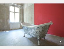 Freistehende Badewanne mit buntem Mosaik auf Löwenfüßen in einem Raum mit roter Wand und verwitterter Putzfassade neben einem großen Fenster mit Rahmen im Retro-Stil.