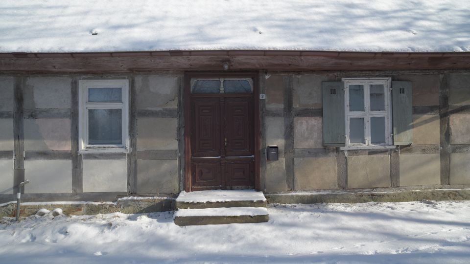 Fassade eines niedrigen Fachwerkhauses mit verschneitem Dach, mittiger dunkler Holztür mit zwei Stufen und zwei Fenstern mit Holzläden. Davor liegt eine schneebedeckte Fläche am Gebäude.