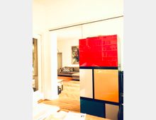 Ein modernes Wohnzimmer mit hellem Holzboden und einer grauen Eckcouch, im Hintergrund ein großformatiges Kunstwerk an der Wand. Im Vordergrund steht ein farbenfroher, geometrisch gestalteter Schrank mit roten, gelben, blauen und weißen Flächen.