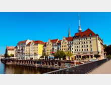 Reihe von historischen Wohn- und Geschäftshäusern mit roten Dächern entlang eines Flusses; im Hintergrund der Fernsehturm und eine Kirchturmspitze. Im Vordergrund eine Promenade und Bäume, darunter ein Bereich mit Graffiti.