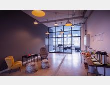 Ein offener, moderner Raum mit Betonboden und minimalistischer Einrichtung, darunter Sessel, Hocker und Tische links sowie eine Kaffeetafel und Snacks rechts. Im Hintergrund befindet sich ein verglaster Konferenzraum mit großen Fenstern, durch die Tageslicht einfällt.