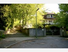 Eingangsbereich eines Wohnhauses mit zwei Wegen: links eine Steintreppe, die von einer Mauer und Bäumen umgeben ist, und rechts ein schmiedeeisernes Tor vor einer Einfahrt. Im Hintergrund befindet sich ein großes Haus mit roten Dachziegeln und mehreren Fenstern.