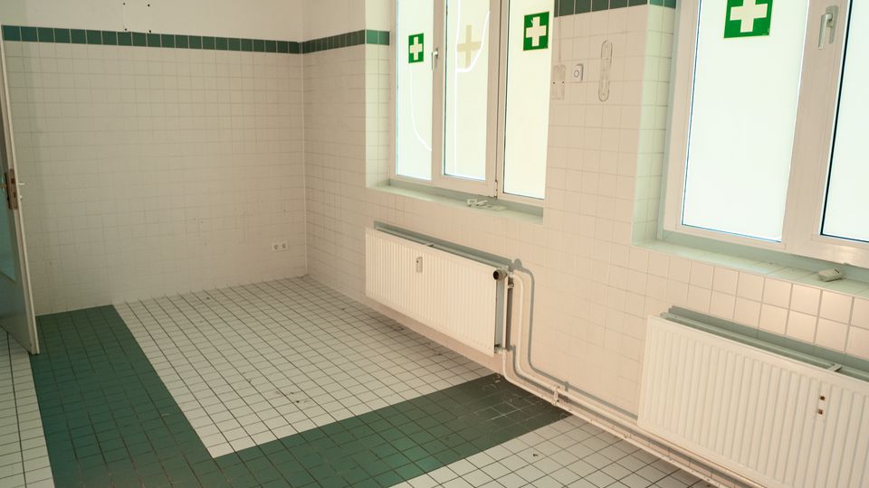 Leerer, gekachelter Raum mit weißer Wandverfliesung und grünem Fliesenstreifen sowie graugrün gemustertem Fliesenboden. An der Fensterwand befinden sich mehrere große Fenster über Heizkörpern; an den Wänden sind grüne Erste-Hilfe-Kreuzsymbole angebracht.