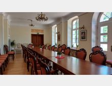 Ein eleganter Konferenzraum mit einer langen hölzernen Tischreihe und gepolsterten Stühlen mit Verzierungen. Große Fenster lassen Tageslicht herein, und die Wände sind mit klassischen Dekoelementen und Gemälden geschmückt.