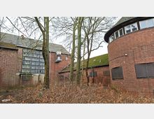 Verlassen wirkender Backstein-Gebäudekomplex mit einem runden Anbau rechts und einem großen Glasbausteinfenster links, mehrere Fenster und Türen sind mit Platten verschlossen. Davor stehen kahle Bäume und dichter Bewuchs auf einem ungepflegten Gelände.