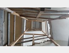 Holzgerüst in einem Gebäudetreppenhaus, mit freiliegenden Wänden aus weißem Putz und teilweise beschädigtem Mauerwerk. Das Gerüst wird verwendet, um Zugang bei Bau- oder Renovierungsarbeiten zu ermöglichen.