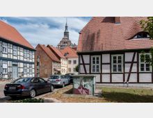 Straßenszene in einer historischen Altstadt mit Fachwerkhäusern, roten Ziegeldächern und geparkten Autos im Vordergrund; im Hintergrund erhebt sich ein Kirchturm mit Spitze.