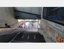 Treppenabgang zu einem Bahnsteig an einem Bahnhof, mit grauen Wänden aus Beton, bedeckt mit Graffiti. Im Hintergrund sind ein Zug, Glasgeländer und Hinweisschilder für Gleise sichtbar, mit Personen, die den Bereich nutzen.