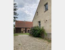 Ein gepflasterter Hof mit zwei Gebäuden; rechts ein verputztes Wohnhaus mit einem Basketballkorb an der Wand und einem blühenden Strauch davor, links ein Fachwerkgebäude mit roten Ziegeln und Holztüren. Der Himmel ist bewölkt.