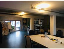 Ein offener, modern gestalteter Büroraum mit einem großen weißen Tisch in der Mitte, umgeben von Bürostühlen. Im Hintergrund sind Regale, eine Glastür mit Blick nach draußen, eine Zimmerpflanze und ein Whiteboard mit Notizzetteln sichtbar.