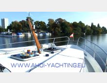 Eine Bootsperspektive auf einem ruhigen Fluss mit einer Anlegestelle und mehreren geparkten Booten im Hintergrund, umgeben von Bäumen im Herbstlaub. Auf dem Deck des Bootes sind Snacks und Getränke arrangiert, und eine kleine rote Flagge ist sichtbar.