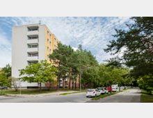 Mehrstöckiges Wohngebäude mit hellgelber und weißer Fassade, umgeben von Bäumen. Davor verläuft eine Straße mit geparkten Autos und einem Gehweg, die Szenerie befindet sich in einem grünen Wohnviertel.