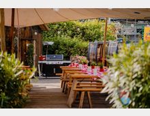 Überdachter Außenbereich mit Holzterrasse und langen Picknicktischen mit rot-weiß karierten Tischdecken und Pflanzendekoration. Im Hintergrund stehen ein großer Gasgrill neben einer Holzwand und eine kleine, verglaste Kabine; Lichterketten und dichtes Grün umgeben den Bereich.