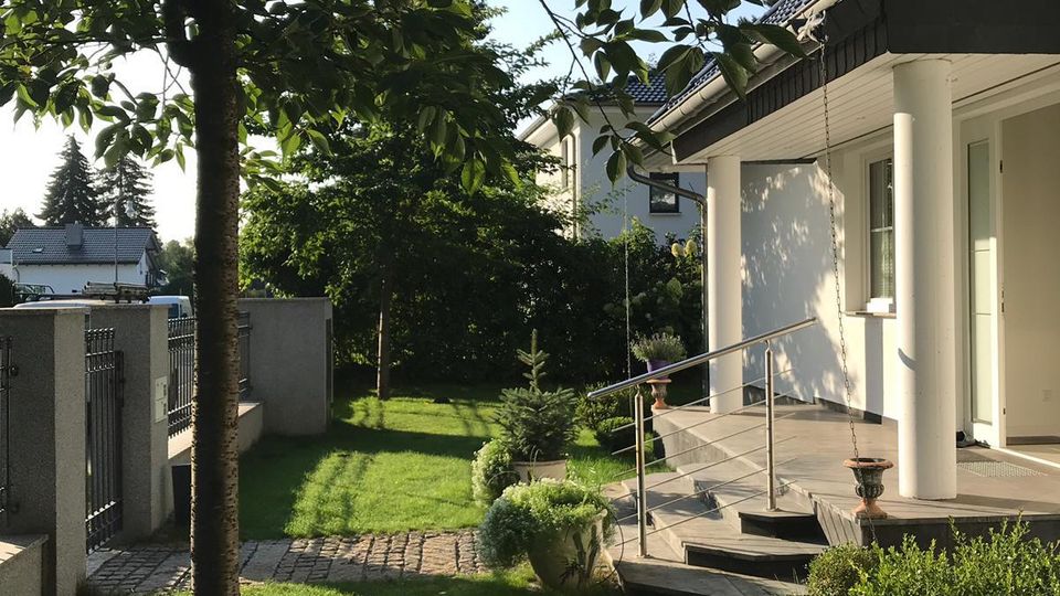Gartenbereich eines Einfamilienhauses mit gepflegtem Rasen, einem Baum im Vordergrund und einem gepflasterten Weg entlang eines Zauns. Rechts ist die weiße Hausfassade mit überdachtem Eingangsbereich, Säulen und Treppe mit Metallgeländer zu sehen.