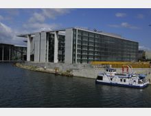 Modernes Bürogebäude mit gläserner Fassade und weißen Betonstreben am Ufer eines Kanals, im Vordergrund ein kleines blau-weißes Ausflugsschiff am Wasser. Im Hintergrund befinden sich Bauarbeiten und weitere moderne Gebäude.