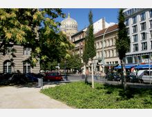 Straßenszene mit einem kleinen Park im Vordergrund, flankiert von historischen und modernen Gebäuden; im Hintergrund ist die goldene Kuppel der Neuen Synagoge zu sehen. Der Bereich ist von Bäumen und parkenden Autos gesäumt, mit Geschäften und Cafés entlang der Straße.