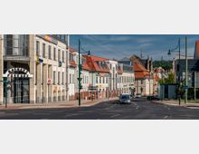 Straßenansicht mit mehrstöckigen Gebäuden und Geschäften, darunter die "Rathaus Passage" mit moderner Glasfassade. Im Hintergrund historische Gebäude mit roten Ziegeldächern und grüner Hügellandschaft.