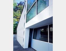 Moderne Gebäudefassade mit glatten Metallpaneelen und großen Glasfenstern, die zum Teil die Reflexion von Bäumen und Himmel zeigen. Im Vordergrund befindet sich ein Kiesboden, und im Hintergrund sind Bäume sichtbar.