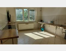 Helle, funktionale Küche mit beigen Fliesen an Wänden und Boden, Holzarbeitsflächen, zwei Spülbecken und einem Herd. Ein großes Fenster mit Blick auf einen Garten sorgt für Tageslicht, und ein Heizkörper befindet sich unter dem Fenster.