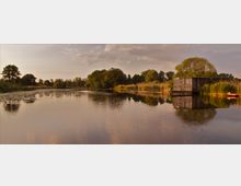 Ein ruhiger See mit dichter Ufervegetation, darunter Schilf und Bäume, im Abendlicht. Am rechten Ufer steht eine einfache Holzhütte, daneben liegt ein rotes Kanu im Gras.