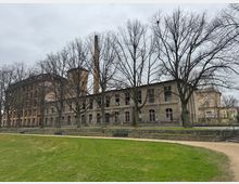 Ehemaliges Fabrikgebäude mit verfallener Fassade aus grauem Mauerwerk und großen, leeren Fenstern, inmitten von kahlen Bäumen. Im Vordergrund eine grasbewachsene Fläche und ein Kiesweg, der entlang einer niedrigen Steinmauer verläuft.