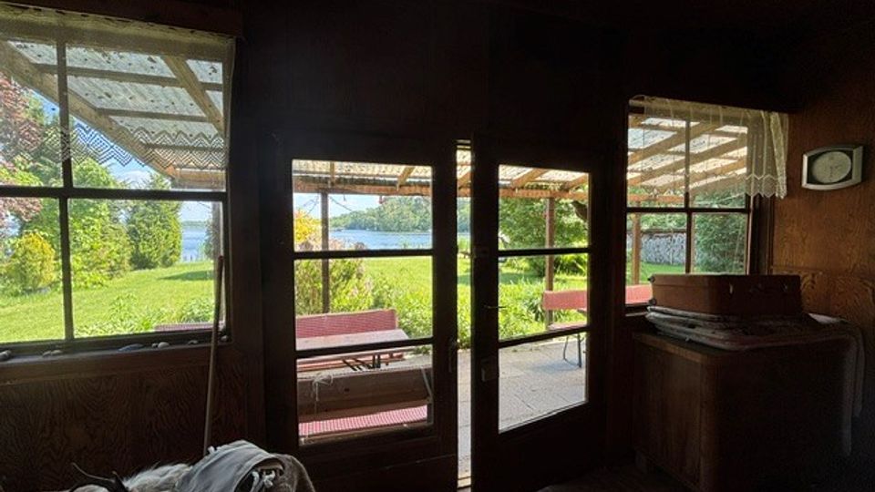 Dunkler Innenraum einer rustikalen Holzhütte mit großen Fenstern und einer Glastür zur überdachten Veranda. Draußen sind grüne Wiese, Sträucher und ein Blick auf ein Gewässer zu sehen, unter dem Vordach stehen rot-weiße Sitzbänke.
