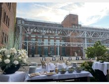 Ein rotes Backsteingebäude im Industrie-Stil mit großen Fenstern und einem Stahlsteg im Vordergrund. Im Vordergrund sind weiß gedeckte Tische mit Blumen, Geschirr und Schüsseln mit Salaten für ein Buffet arrangiert.