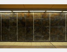 U-Bahn-Station mit einer Wand aus dunkel glänzenden Fliesen mit bronzefarbenen und schwarzen Mustern. Der Boden besteht aus beige und braun gefliesten Bereichen, während eine gelbe Decke und Deckenbeleuchtung den Raum beleuchten.