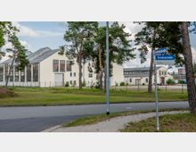 Industriegebiet mit weißen Lagerhallen und großen Fenstern, umgeben von einem Zaun, Bäumen und einer Wiese. Im Vordergrund befindet sich eine Straßenkreuzung mit Schildern für die "Fabrikenstraße" und "Grisutenstraße".