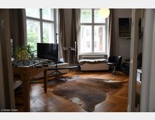 Wohnzimmer mit großen Fenstern, die von beigefarbenen Vorhängen umrahmt sind. Der Raum enthält einen Fernseher, einen Kicker, eine Tischlampe, einen schwarzen Stuhl, ein weißes Sofa und einen Teppich in Felloptik auf Holzboden.