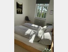 Helles Schlafzimmer mit einem Holzbett, das mit grau-weiß gestreifter Bettwäsche bezogen ist. Im Hintergrund ein großes Fenster mit Blick ins Grüne, ein Stuhl, ein kleines Beistelltischchen und ein an der Wand hängendes Bild.