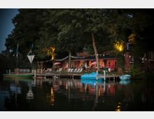 Backstein-Gebäude an einem Gewässer, umgeben von Bäumen, mit einer beleuchteten Terrasse, Liegestühlen und dekorativen Wimpelketten; im Vordergrund befinden sich ein Steg und blaue sowie grüne Boote auf dem Wasser.