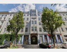 Straßenansicht eines mehrstöckigen Wohngebäudes mit hellem Putz und dekorativen Elementen an der Fassade. Im Erdgeschoss befinden sich Geschäfte mit blauen Fliesen und Graffitis, und ein Torbogen führt zu einem Innenhof; davor stehen mehrere Bäume und parkende Autos.